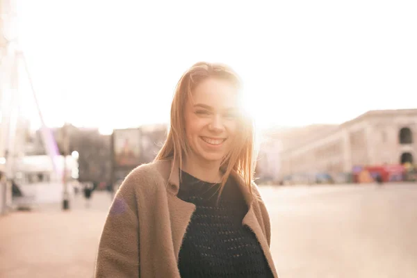 Happy girl in casual wear,wearing a coat,standing in the background of the street landscape in the sunset,looking in camera and smiling.Portrait of a beautiful lady on the street, posing on the camera