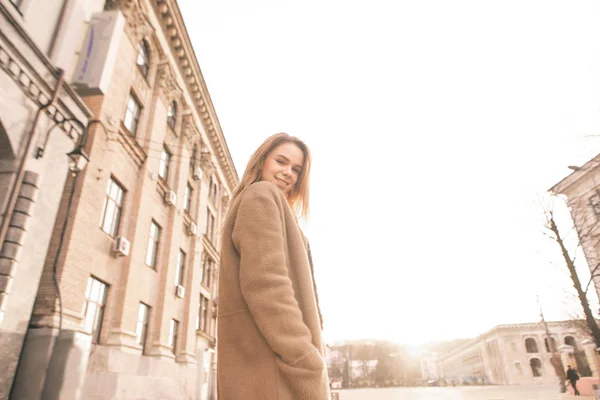 Ευτυχισμένο κορίτσι σε περιστασιακή ένδυση, φορώντας ένα παλτό, στέκεται στο φόντο του τοπίου του δρόμου στο ηλιοβασίλεμα, κοιτάζοντας στην κάμερα και χαμογελώντας. Πορτρέτο μιας όμορφης κυρίας στο δρόμο, ποζάρει στην κάμερα — Φωτογραφία Αρχείου