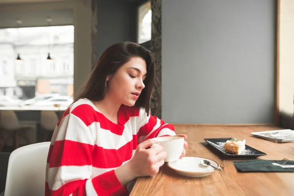 Приваблива брюнетка носить повсякденний одяг, сидячи в кафе біля вікна з кавою та десертом. Красива дівчина п'є каву в кафе . — стокове фото