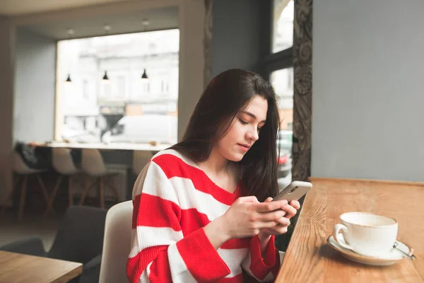 Красива брюнетка дівчина використовує смартфон у кафе, сидячи за столом з чашкою кави та десертом. Дівчина-підліток пише повідомлення на смартфоні в кафе . — стокове фото