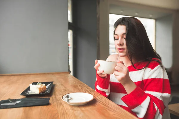 Menina bonita em um vestido casual sentado em um café, segurando uma xícara de café em suas mãos e apreciando o cheiro. Menina atraente beber café em um café acolhedor — Fotografia de Stock