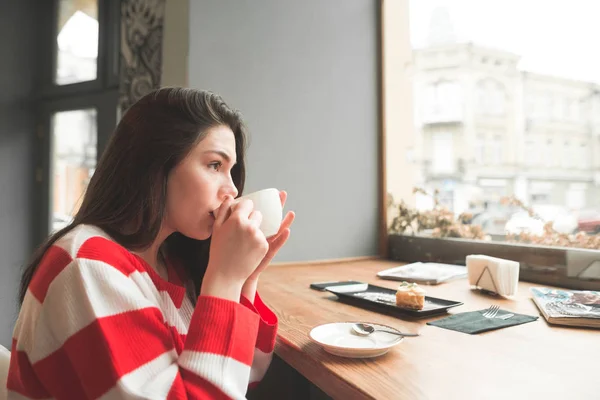 Linda chica bebe café caliente en un acogedor café, mirando por la ventana. Hermosa chica platos de café y postre en el restaurante . — Foto de Stock