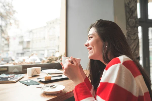 Menina feliz sentada à mesa em um café com uma sobremesa e uma xícara de café delicioso em suas mãos, olhando para a janela e sorrindo. — Fotografia de Stock