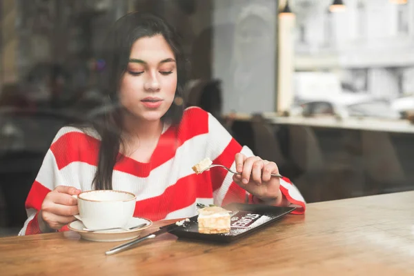 Κορίτσι κάθεται σε ένα ζεστό καφέ στο τραπέζι κοντά στο παράθυρο, τρώγοντας ένα ορεκτικό επιδόρπιο και κρατώντας ένα φλιτζάνι καφέ. Ελκυστικό κορίτσι τρώει τούρτα σε ένα εστιατόριο και πίνοντας καφέ, κοιτάζοντας ένα επιδόρπιο — Φωτογραφία Αρχείου