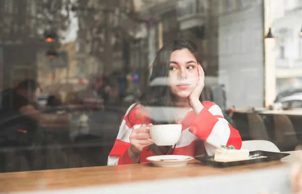 Симпатичная девушка в ярком повседневном платье сидит в кафе у окна с чашкой кофе и тарелкой десерта на столе и смотрит в сторону. Copyspace — стоковое фото