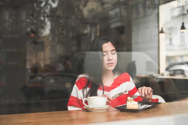 Милая девушка в повседневной одежде отдыхает в кафе с чашкой кофе и десертом в выходной. Портрет девушки в кафе через окно ресторана, смотрящей на тарелку с десертом — стоковое фото