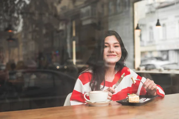 Позитивная привлекательная девушка носит яркое повседневное платье, сидит в кафе с тарелкой десерта и чашкой кофе, держит в руках вилку, смотрит в камеру и улыбается. Copyspace — стоковое фото