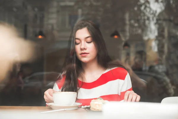 Retrato de una adolescente sentada en una mesa en el café con una taza de café y un plato de postre, foto a través de la ventana desde la calle.Atractiva morena en el café en la mesa cerca de la ventana — Foto de Stock