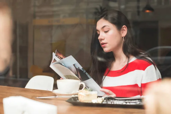 Söt brunett flicka sitter i ett kafé med en kopp kaffe och en dessert på bordet, håller en tidning och läser i hennes händer. Porträtt av en flicka som vilar i ett kafé. — Stockfoto