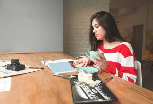Attraktiv flicka sitter vid ett bord i ett kafé med en tallrik dessert, och en kopp kaffe i händerna, läser noggrant tidningen. Fritid i caféet för en kopp kaffe och läsning. — Stockfoto