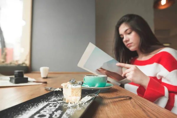 Привлекательная девушка сидит за столом в кафе с тарелкой десерта и чашкой кофе в руках, внимательно читает журнал. Отдых в кафе на чашечку кофе и чтение . — стоковое фото