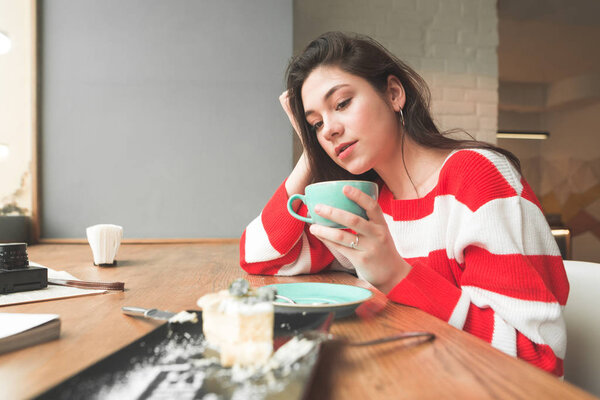 Attractive brunette girl sits in a cafe with a cup in her hands and looks at a plate of dessert. Girl dishes with cake and coffee in a cozy cafe.