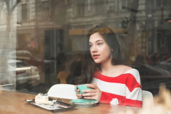 Vacker flicka tillbringar tid i ett kafé, sitter vid ett bord med en tallrik dessert och en kopp kaffe i hennes händer, ser tankfullt åt sidan. Brunett rätter i ett kafé. — Stockfoto