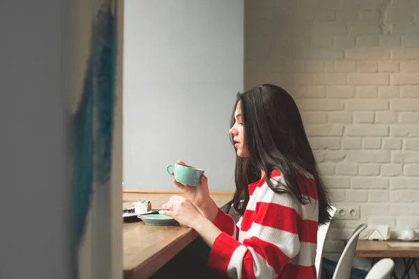 Привлекательная брюнетка носит повседневную одежду, сидя в кафе рядом с окном с кофе и десертом. Красивая девушка пьет кофе в кафе и ест десерт. Вид сбоку — стоковое фото