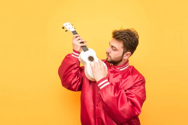 Homem de casaco vermelho toca guitarra havaiana no fundo de uma parede amarela. Estudante com barba é isolado em um fundo amarelo com um ukulele na mão. Conceito musical — Fotografia de Stock