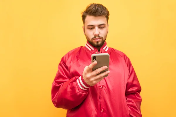 Hombre barbudo con una chaqueta roja lleva un teléfono inteligente sobre un fondo amarillo, se centra en la pantalla del teléfono. Concéntrate en el smartphone. Aislado . —  Fotos de Stock