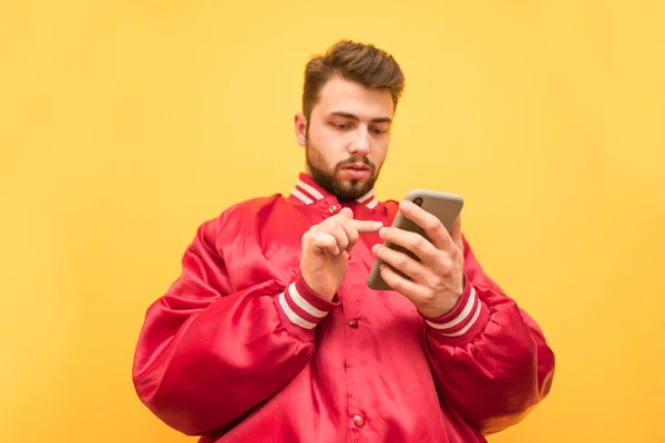 Hombre adulto con barba lleva una chaqueta roja, se coloca sobre un fondo amarillo y utiliza un teléfono inteligente, hace clic en la pantalla con el dedo, boca abajo en la pantalla. Aislado . —  Fotos de Stock