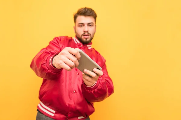赤いジャケットとあごひげを着た男は、スマートフォンに焦点を当てて、スマートフォンでビデオゲームをすることに集中しています。黄色の背景に分離されています。モバイルゲーム. — ストック写真