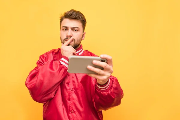 Retrato de un hombre serio con barba, con una chaqueta roja, de pie con un teléfono inteligente en la mano sobre un fondo amarillo y mira cuidadosamente a la pantalla.Juego enfocado está jugando juegos móviles . — Foto de Stock