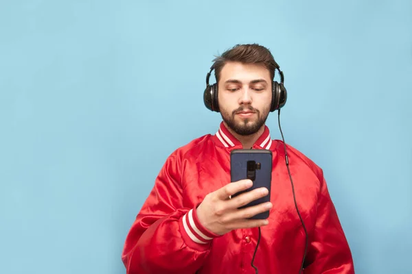 Портрет бородатого мужчины в наушниках, стоящего на фоне синей стены со смартфоном в руке, смотрящего сфокусированным на экране в красной куртке. Isolated . — стоковое фото