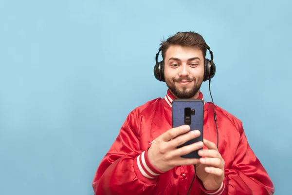 Retrato de un hombre positivo con barba, escucha música en auriculares y usa un teléfono inteligente sobre un fondo azul, con una chaqueta roja. Aislado . —  Fotos de Stock