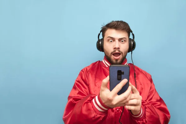 Смешной человек в красной куртке и с бородой стоит на синем фоне с наушниками и смартфоном в руках, с шокирующим лицом, смотрящим на экран телефона в красной куртке. Isolated . — стоковое фото
