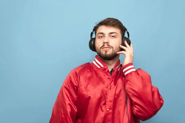 Retrato de um homem barbudo em seus fones de ouvido e um casaco vermelho em um fundo azul, olhando para a câmera. Homem adulto ouve música em fones de ouvido. Isolados . — Fotografia de Stock