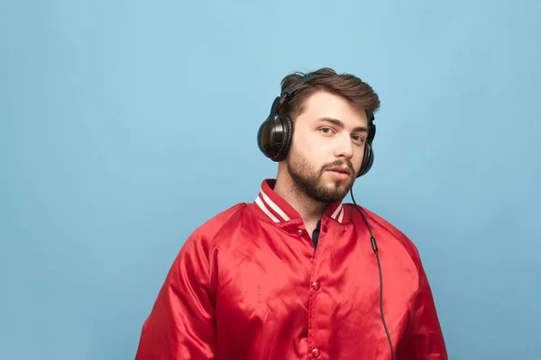 Retrato de um homem com barba, vestindo uma jaqueta vermelha, fica no fundo de uma parede azul, e ele ouve música nos fones de ouvido. . — Fotografia de Stock