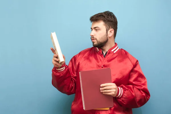 Portret amerykańskiego studenta z książek i notebooków w rękach ubrany w czerwoną kurtkę, odizolowany na niebieskim tle. Nauka koncepcji. Portret mężczyzny z brodą i książek w ręku — Zdjęcie stockowe