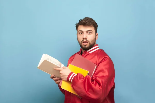 Amazed young man with a beard and books in his hands, wearing a red jacket, looks into the camera with shocking face. Portrait of astonished student husband. Studying at the University of Concepts. — Stock Photo, Image