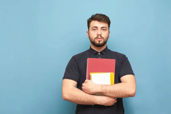 Портрет ученика с бородой, в темной рубашке, стоящего на синем фоне с книгами и блокнотами в руках и смотрящего в камеру с серьезным лицом. Isolated . — стоковое фото