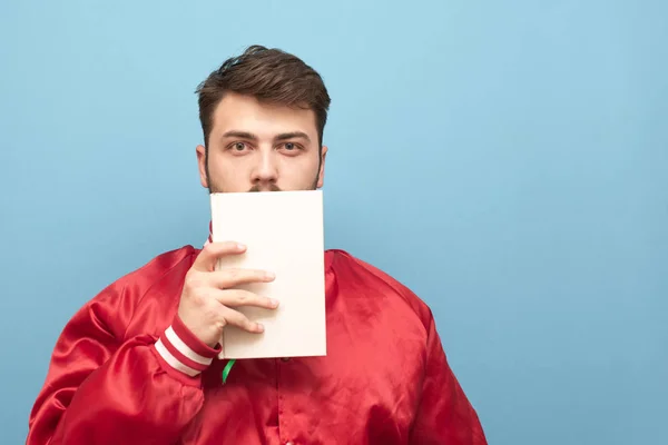 Homem adulto em uma jaqueta vermelha fica em um fundo azul com um livro branco na mão e olha para a câmera. Retrato estudantil segura o livro em torno de seu rosto. Isolado. Espaço de cópia — Fotografia de Stock