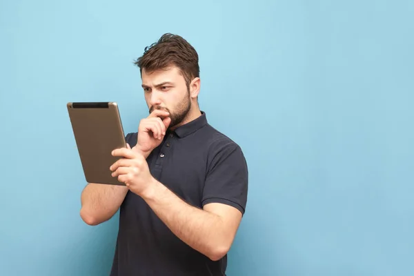 Un hombre concentrado con barba y camiseta está de pie sobre un fondo azul y mira la pantalla con cuidado, vistiendo una camiseta oscura. Aislado. Hombre de negocios utiliza una tableta . — Foto de Stock