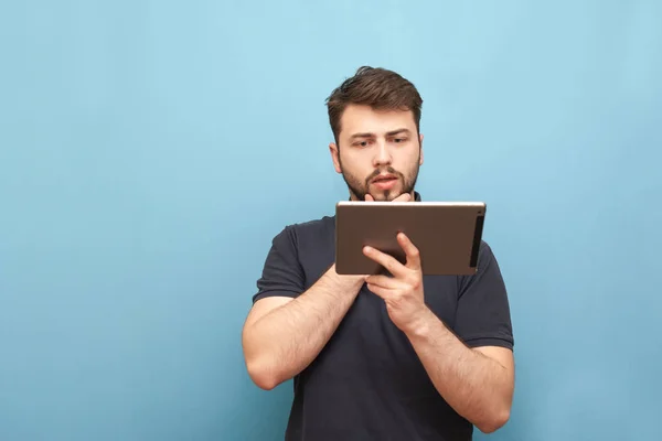 Porträt eines Studenten mit Bart, der auf blauem Grund steht und das Internet auf seinem Smartphone nutzt, es sorgfältig vom Tablet liest und lernt. Mann mit Tablette in der Hand, isoliert. — Stockfoto