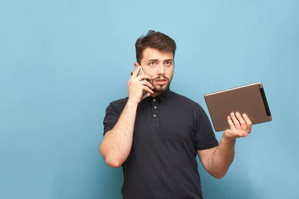 Portrait of a busy man standing on a blue background with a tablet in his hands and talking on the phone.Business man with gadgets in his hands is isolated on a blue background. Copyspace