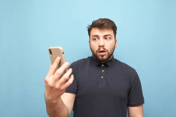 El hombre asombrado con barba se para con un smartphone en las manos y parece confundido con una cara confusa. Divertido hombre sostiene un teléfono inteligente en sus manos, aislado sobre un fondo azul — Foto de Stock