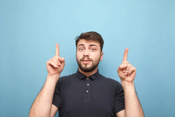 Выразительный мужчина с бородой, стоящий на синем фоне, глядя в камеру с изумленным лицом и показывая свои пальцы в пустом месте, в темной футболке — стоковое фото