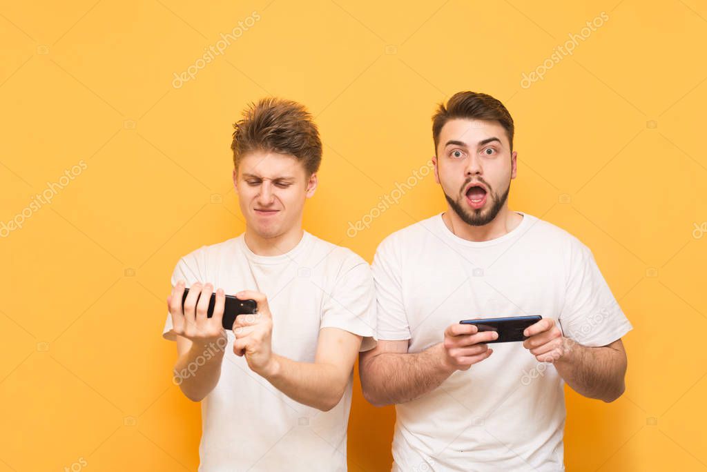 Emotional gamers play mobile games on a smartphone, wear white t-shirts, one looks at the camera, the other in the smartphone screen, they are isolated on a yellow background.