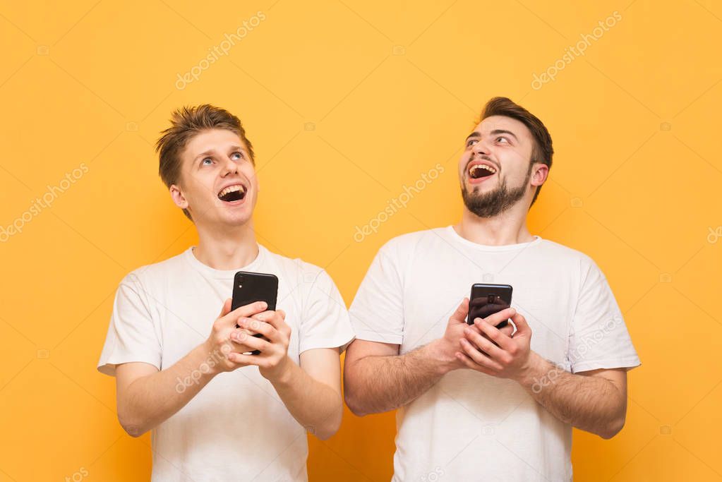 Two smiling men wearing a white T-shirt are standing with smartphones in their hands on a yellow background and looking up. Two happy people hold a smartphone and look at the place for advertising