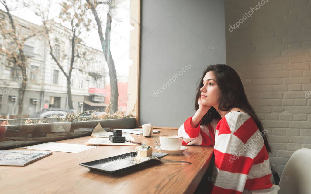 Attractive girl, bright clothes, sitting in a cafe with a plate of dessert and a cup of coffee, looks dreamily in the window.Cute lady sits in a cafe for a thoughtfully looking into the window.