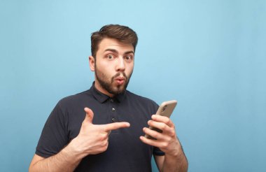 Karanlık bir T-shirt ve sakal hayran adam, elinde bir akıllı telefon tutar, kamera duygusal görünüyor ve telefonda işaret parmağı gösterir. Elinde akıllı telefon olan duygusal adam. Izole.