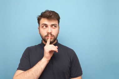 Bir sakalı ile yetişkin bir adamın portresi, boş bir yere yakın bir şekilde bakıyor ve mavi bir arka planda ağzında bir parmağını tutan bir sessizlik belirtisi gösteriyor. Izole.