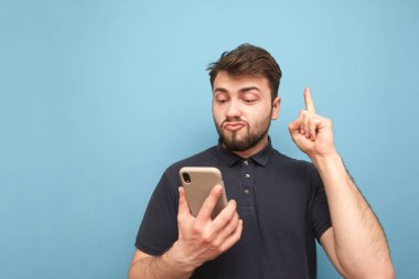 Sakal tutan duygusal adam, parmaklarını boş bir uzayda gösteren bir akıllı telefonu elinde tutuyor. Karanlık bir tişört şaşırttı yetişkin bir akıllı telefon kullanır ve copyspace bir parmak gösterir. Mavi arka plan