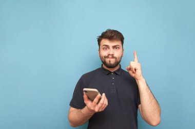 Mavi bir arka planda bir sakal ve elinde bir telefon ile komik bir adam portre, kamera bakarak ve komik bir yüz yapma. Bir smartphone ile şaşkın adam boş bir alan gösterir. Izole.