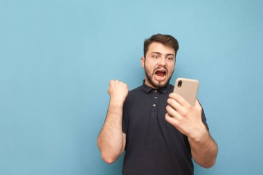Olumlu genç adam elinde bir akıllı telefon tutar, ekrana bakar ve elleri ile sevinler kaldırdı. Adam, mavi bir arka planda izole başarı gülümsüyor ve seviniyor. Boşaltmak