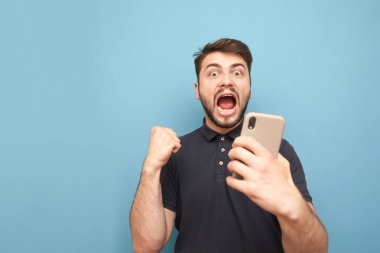 Duygusal sakallı adam elinde Smartphone tutar, kamera ve sevinler bakıyor. Elinde bir akıllı telefon ile adam başarı, mavi bir arka planda izole sevinç. Boşaltmak