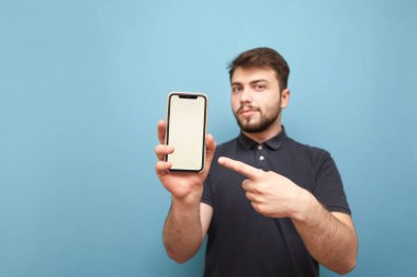 Gülümseyen adam elinde bir smartphone tutar ve mavi bir arka planda beyaz bir ekranda parmağını gösterir. Sakallı adam koyu bir T-shirt giyer ve kamera beyaz bir ekran ile bir akıllı telefon gösterir.