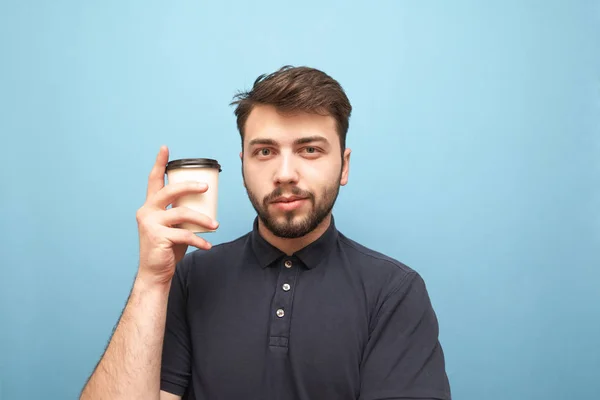 Close-up portrait of a man with a beard standing on a blue background with a paper cup of coffee in his hands, looks into the camera with a face-to-face. Isolated.