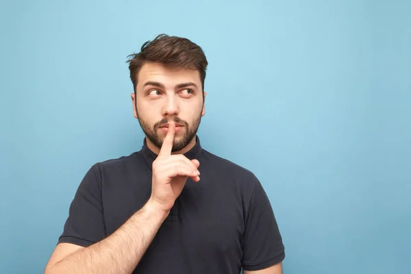 Portrait of an adult man with a beard thoughtfully looking sideways to a blank place and showing a sign of silence holding a finger near his mouth on a blue background. Isolated.