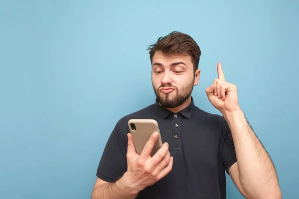 Emotional man with a beard holds a smartphone in his hands, showing his finger up in a blank space. Surprised adult in a dark T-shirt uses a smartphone and shows a finger on copyspace.Blue background — Stock Photo, Image